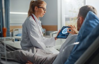 Hospital Ward: Friendly Female Doctor Talks to Sick Male Patient Resting in Bed, Uses Tablet Computer, Shows and Explains Brain Scan Results. Physician Talks to a Man Recovering after Surgery